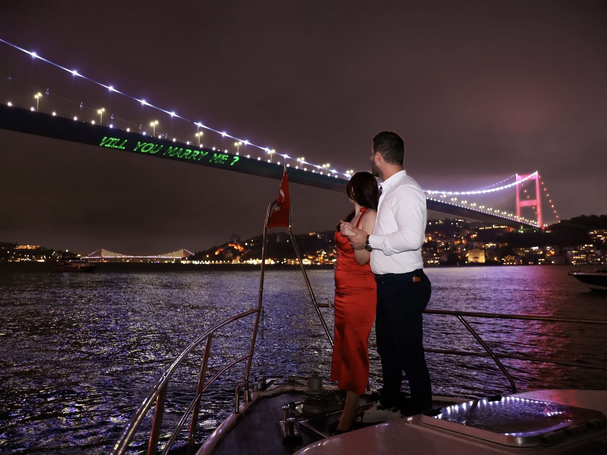 Paar am Bug der Yacht, an der Bosporus-Brücke die Laserschrift Will you marry me