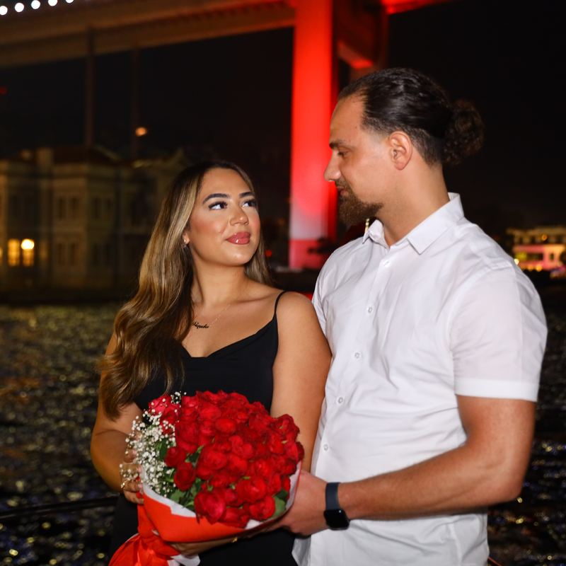 Couple with a bouquet of roses on deck in front of the red-lit Bosphorus Bridge